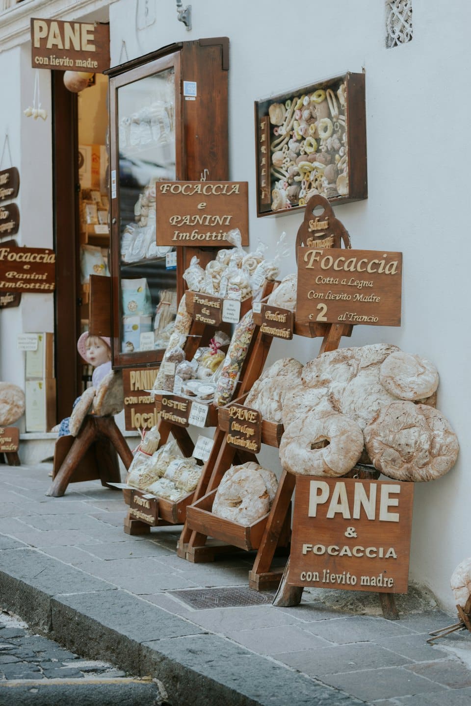 Un bambino seduto vicino alla vetrina di un panificio italiano con pane fresco in esposizione.